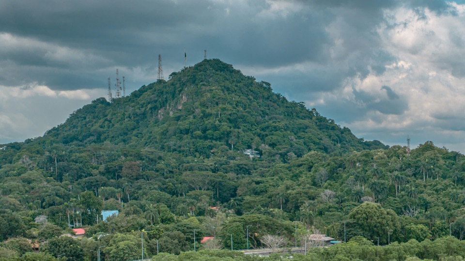 El Cerro Ancón cubierto de bosque, con la bandera y las antenas en la cima, bajo nubes de tormenta